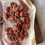 Baked spiced pecans on a sheet of parchment that is resting on a wooden serving board.