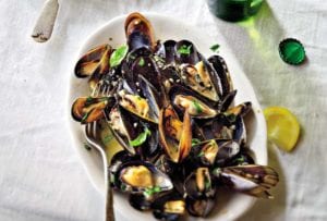 White plate with steamed mussels in beer, topped with parsley, nearby lemon wedges and a bottle of beer