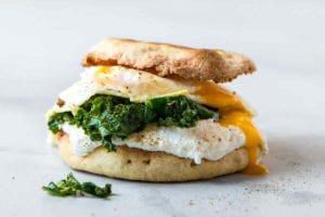 A breakfast sandwich with fried egg, kale, and ricotta sandwiched between two toasted English muffin halves.