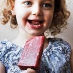 A little girl eating a strawberry ice pop.