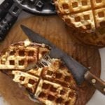 Two waffle iron grilled cheese sandwiches on a wooden cutting board with a knife and a waffle iron nearby.