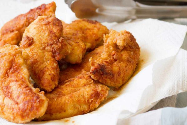Eight pan-fried chicken tenders piled on paper towels on a plate.