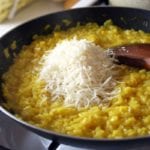 Skillet on the stove filled with risotto alla Milanese, a pile of cheese, and a wooden spoon