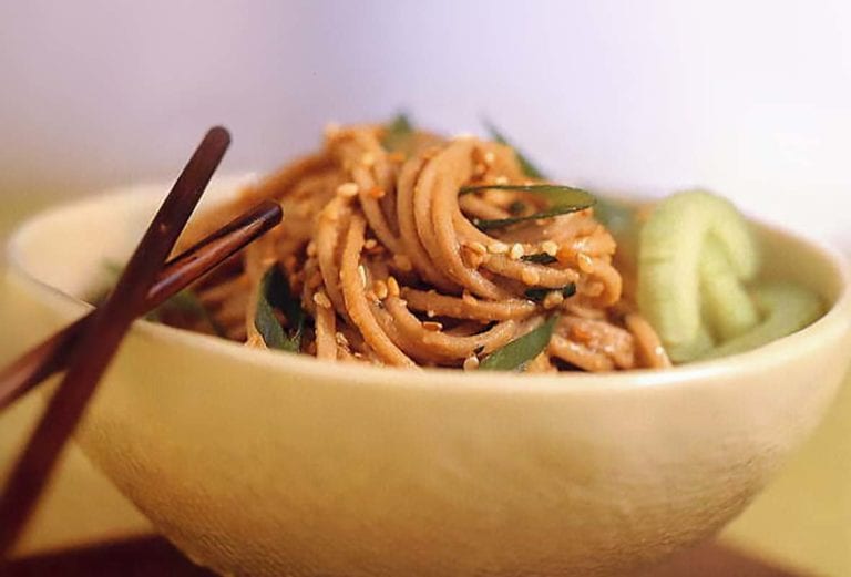 A bowl of sesame-peanut noodles coated in peanut sauce, topped with sesame seeds, chopsticks on the side