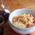 A white bowl filled with farfelle with fresh salmon is nested inside another white bowl with a carafe and glass of red wine in the background.