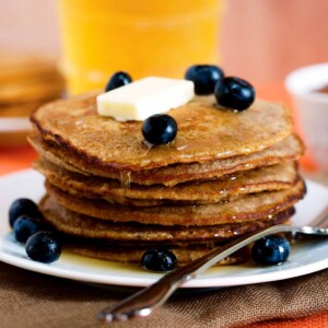 Gingerbread pancakes stacked on a plate, covered with syrup, blueberries, and a pat of butter.