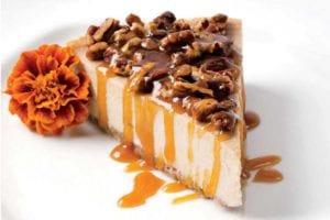A slice of banana rum cheesecake with maple rum sauce and pecan pieces on top and a marigold beside the cake.