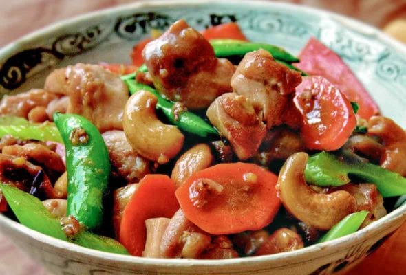Bowl of cashew chicken with chunks of stir-fried chicken, carrots, sugar snap peas, celery, cashews