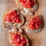 Four slices of bruschetta topped with tomato on a wooden cutting board.