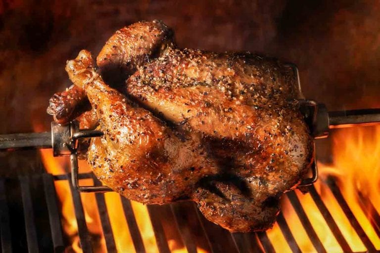 A rotisserie chicken suspended over a grill with flames below.
