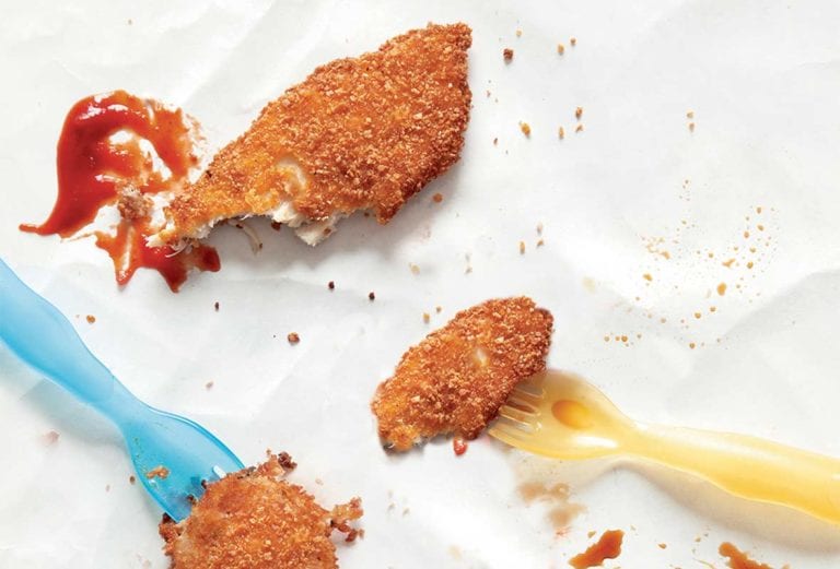 Pieces of chicken fingers, a smear of ketchup and two toddler sized forks on a white sheet.