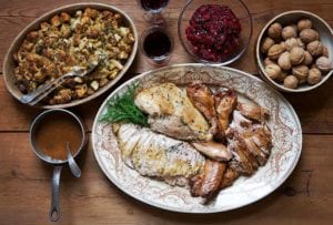 An oval platter with roasted and braised turkey, a dish of stuffing, a bowl of cranberry sauce, a bowl of chestnuts, two glasses of wine, and a small pot of gravy on a wooden table.