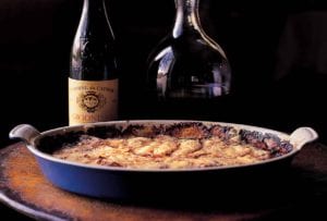 A blue gratin dish filled with potato gratin and a two wine bottles in the background