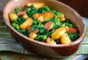 Pieces of sauteed broccoli rabe and slices of seared potatoes in a brown oval dish on a green bench