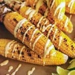 Six ears of grilled Indian corn drizzled with curry yogurt and lime wedges on the side.