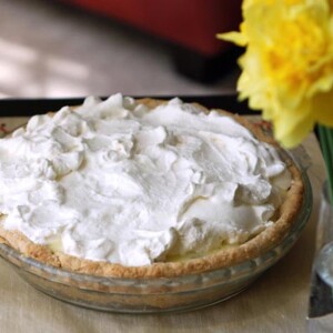A banana cream pie, topped with pillowy meringue in a glass pie dish with a vase of daffodils beside it.