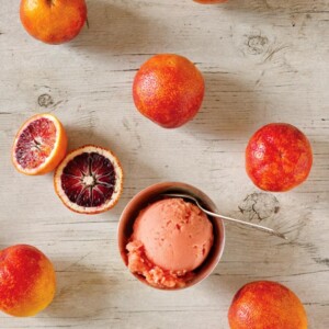 A bowl with a scoop of blood orange sorbet and a spoon in it, surrounded by several blood oranges.