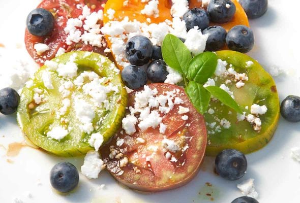 Sliced tomatoes with blueberries and crumbled feta scattered on top and a sprig of basil for garnish.