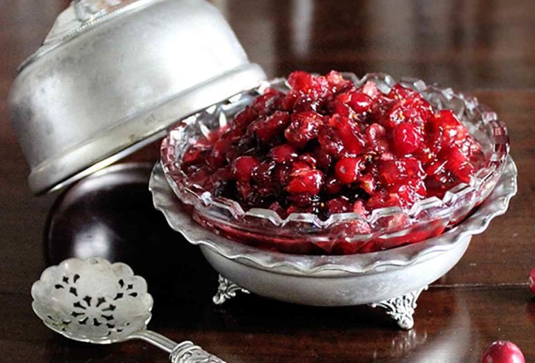 A silver and glass decorative dish filled with Raw Cranberry Sauce. A lid for the dish and a silver spoon rest beside the dish.