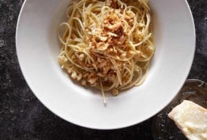 A white bowl filled with spaghetti topped with bread crumbs and a chunk of pecorino beside the bowl.