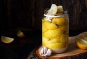 A jar of preserved lemons on a wooden board with lemon wedges and a few garlic cloves beside it.
