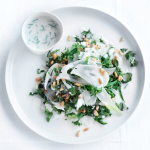 A white plate of kale slaw with shaved fennel and toasted pine nuts on top and a bowl of buttermilk dressing on the side.