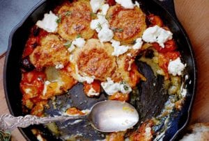 A cast-iron skillet half-filled with tomato and goat cheese cobbler and a spoon resting in the skillet.