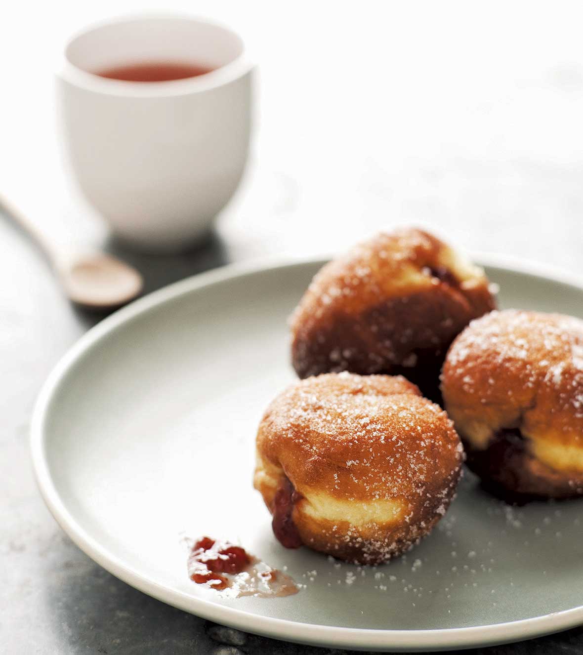 Three German Berliners, or jelly doughnuts, with raspberry jam spilling out on a plate with a cup of coffee
