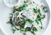 A white plate of kale slaw with shaved fennel and toasted pine nuts on top and a bowl of buttermilk dressing on the side.
