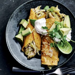 Two chicken enchiladas with green sauce in a black shallow bowl with cubed avocado, sour cream, cilantro, and a lime half.