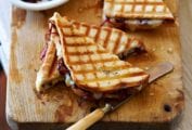 Two halved grilled cheese with onion jam on a wooden cutting board alongside a dish of onion jam and a butter knife.