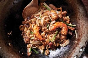 A metal wok with a serving of char kway teow and a wooden spoon resting inside.
