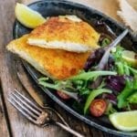 A halved pork Milanese cutlet on a grey oval plate with a lemon wedge and mixed spring greens.