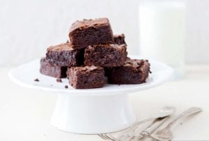 A white cake stand of a pile of one-pot cocoa brownies, a glass of milk, on a white table
