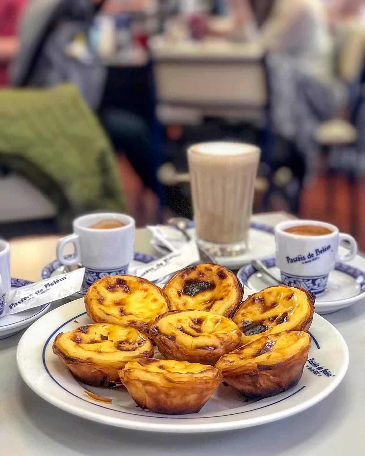 Seven pasteis de Belem, or Portuguese custard pastries, on a plate, with coffee cups nearby