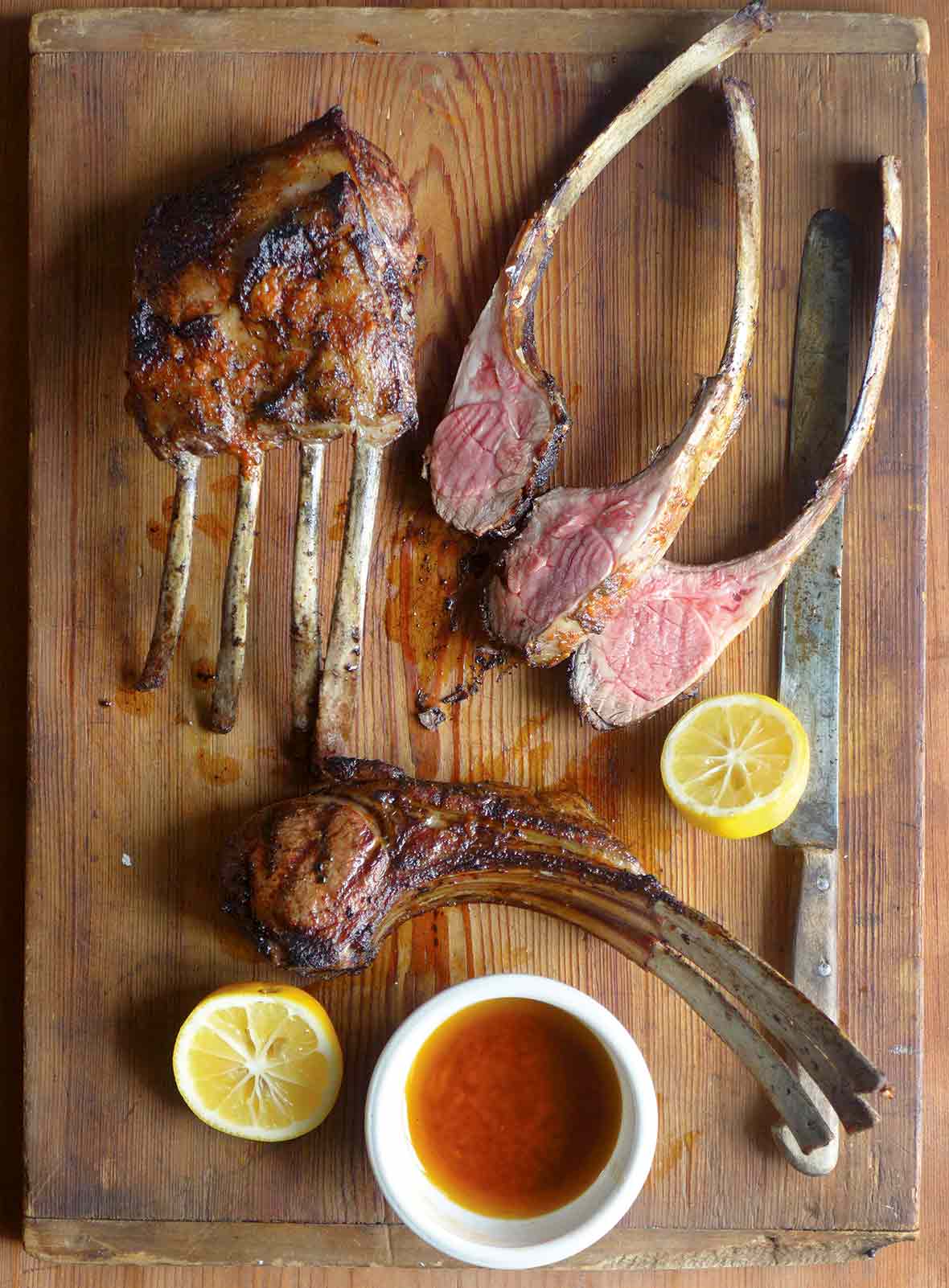 A cutting board with lamb chops, some in racks, some sliced, along jus and lemon halves
