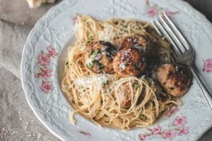 A patterned bowl filled with turkey meatballs with angel hair pasta, and a piece of bread and glass of white wine next to the bowl.