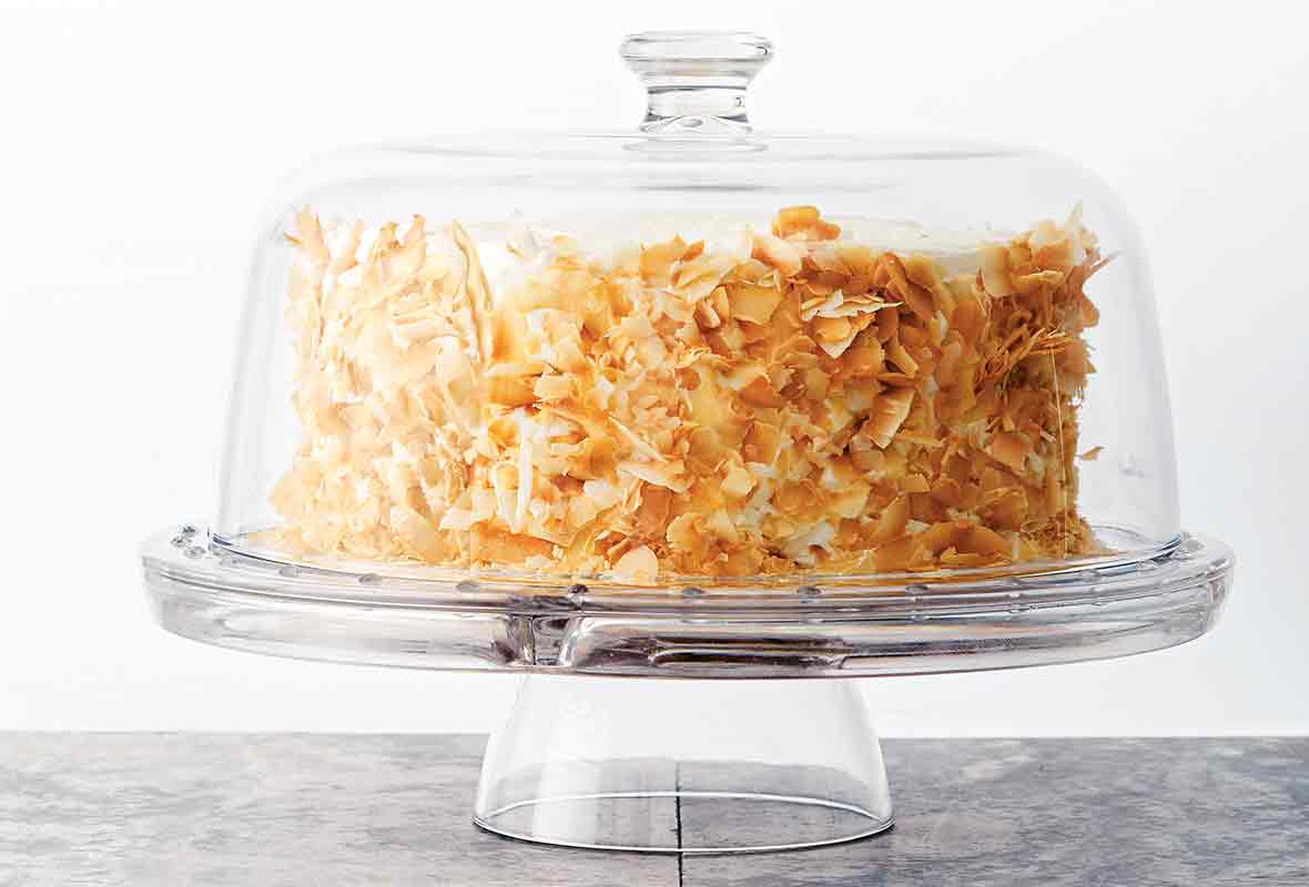 A coconut carrot cake with cream cheese frosting and toasted coconut on the sides on a glass cake platter with a glass domed lid.