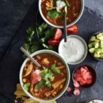 Two bowls of chicken posole, with a bowl of sour cream, some tomato wedges, cilantro, sliced radish, and tortilla shells scattered around them.