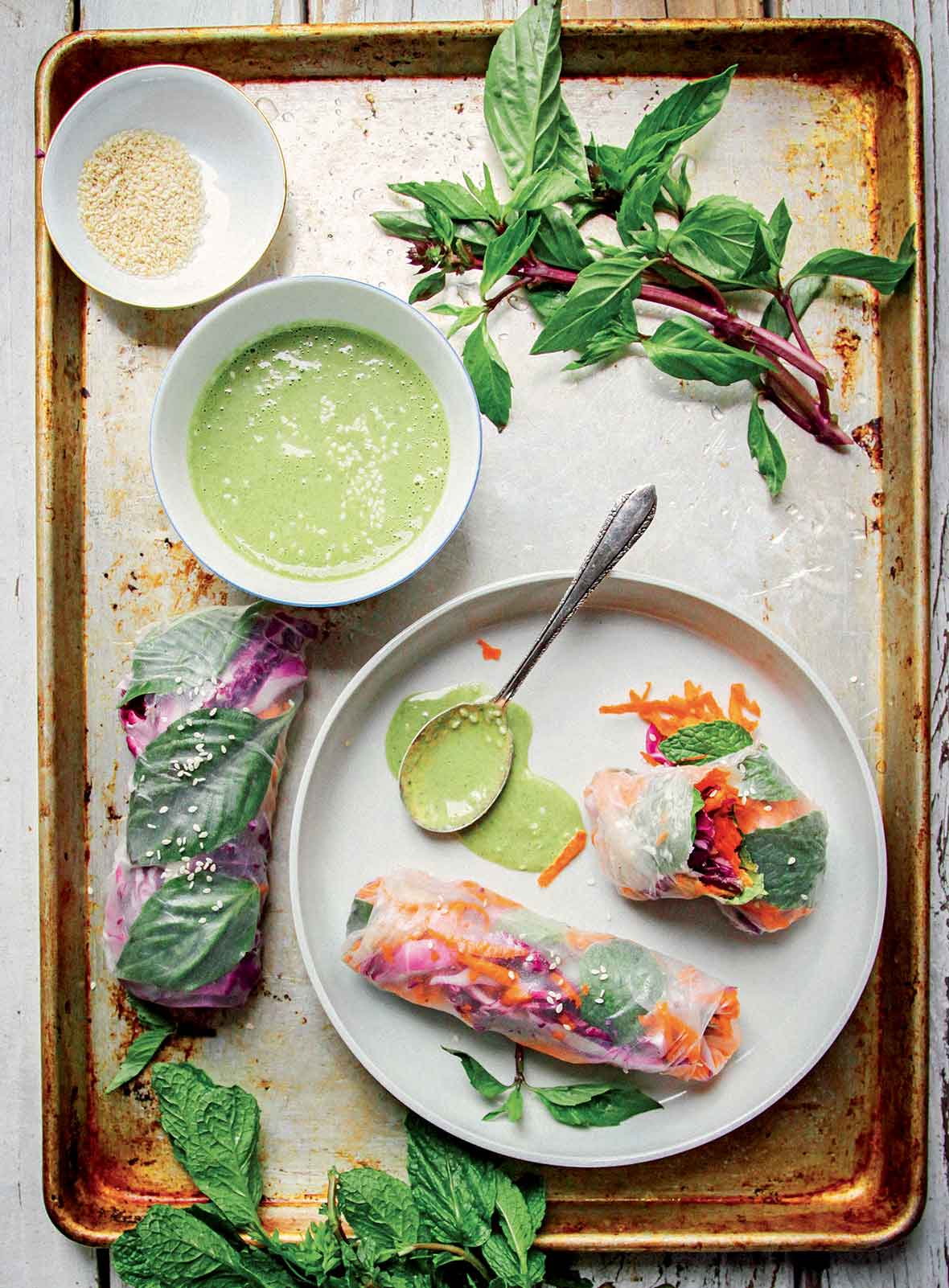 A metal sheet pan with a white plate of summer rolls, a bowl of dressing, and a bowl of sesame seeds, garnishes of Thai basil.