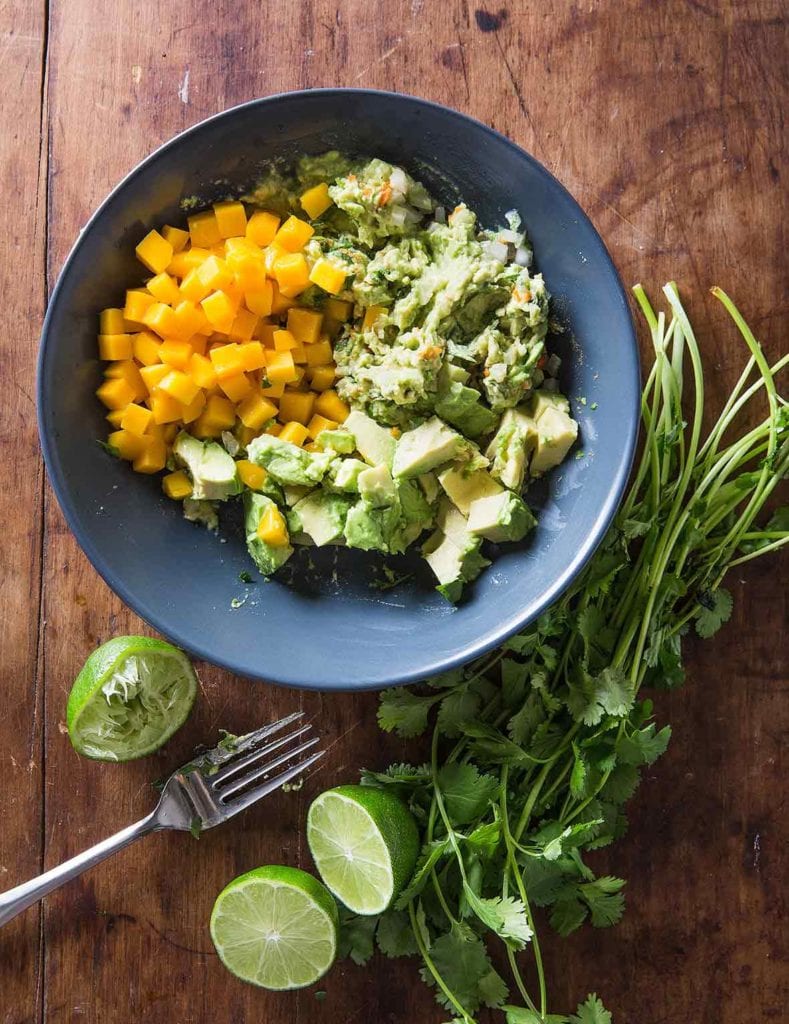 A blue bowl filled with the components of mango guacamole, with a bunch of cilantro, halved limes, and a fork on the side.