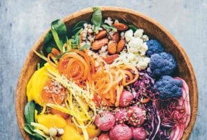 A wooden bowl filled with shredded carrots and red cabbage, purple cauliflower, radishes, broccoli, almonds, and rice