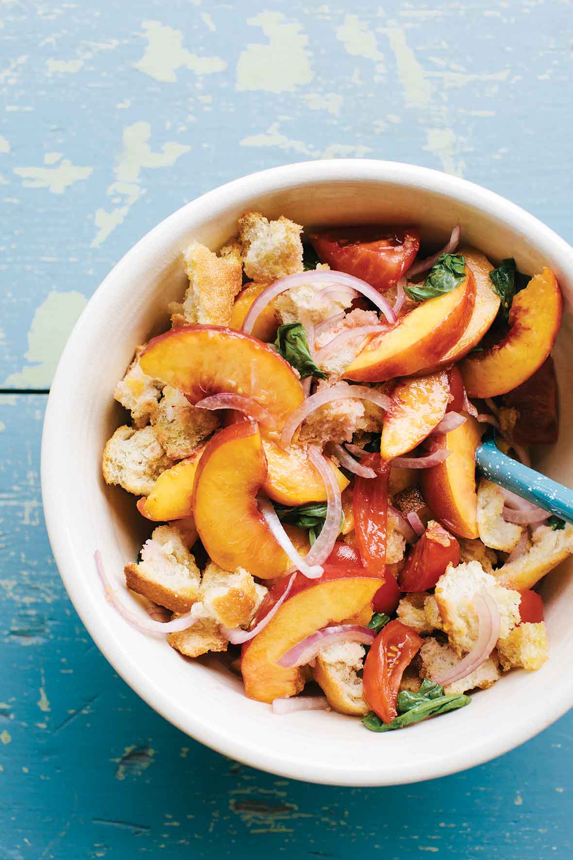 A white bowl filled with tomato and peach panzanella on a blue wooden surface.