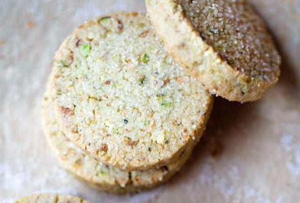 Five round pistachio shortbread cookies with a few broken pistachios on the side
