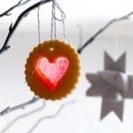 A scalloped round sugar cookie with a heart-shaped windowpane cutout hanging form a tree branch