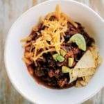 White bowl of beef chili with grated cheese, tortilla chips, and lime wedges on wood
