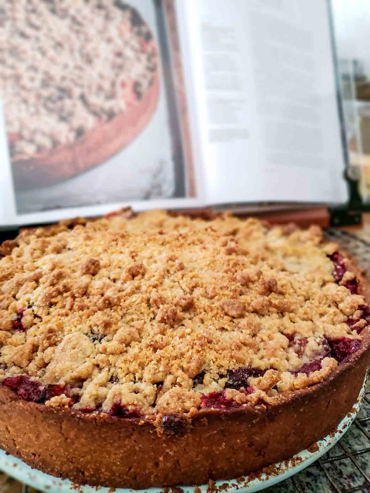 An apricot tart with a thin crust and a crumble topping on a plate in front of an open cookbook showing the same tart