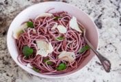 A white bowl of red wine spaghetti with shaved Parmesan and basil leaves.