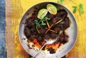 A stainless steel skillet filled with beef rendang and topped with two lime halves, lemongrass, and cilantro