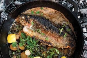 A grilled whole trout in a cast-iron skillet with sliced potatoes, parsley, and lemon